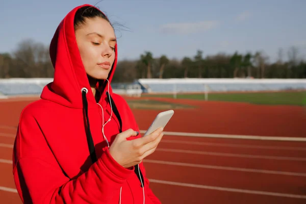 Vista lateral de una joven deportista con una sudadera con capucha roja. Corre y escucha música con su smartphone en las manos . — Foto de Stock