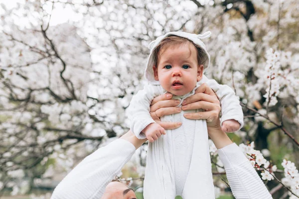 Портрет счастливого радостного ребенка в белой одежде на фоне цветущих деревьев. Семья играет вместе на улице. Мама весело держать маленькую дочь — стоковое фото