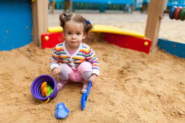 Ein kleines Mädchen mit zwei Schwänzen in einer gestreiften bunten Jacke spielt im Sandkasten auf dem Spielplatz — Stockfoto