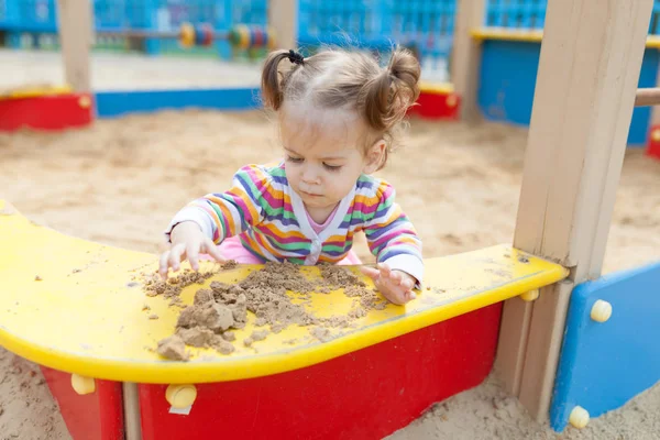 Ein kleines Mädchen mit zwei Schwänzen in einer gestreiften bunten Jacke spielt im Sandkasten auf dem Spielplatz — Stockfoto