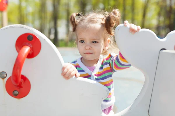 Een klein meisje met twee staarten is gekleed in een gestreepte kleurrijk jasje is spelen op de speelplaats — Stockfoto