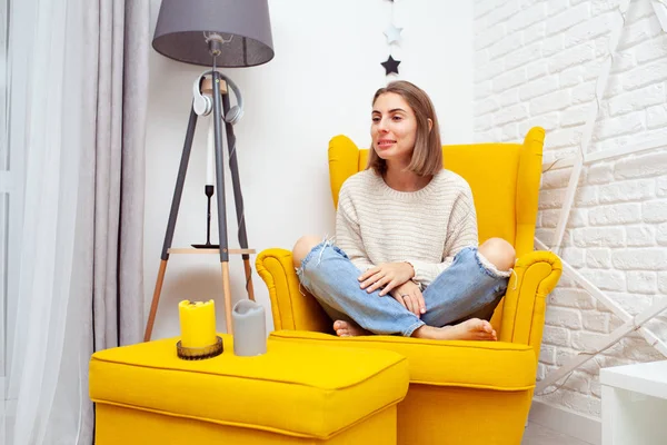 Evde boş zamanların tadını çıkarıyorum. Yumuşak sarı bir sandalyede dinlenen güzel genç bir kadın. Aromatik mumlar yan tarafta duruyor. — Stok fotoğraf