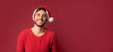 Kırmızı kazaklı ve Noel Baba şapkalı genç yakışıklı adam stüdyoda kırmızı arka planda duruyor ve dişleri gülümsüyor.