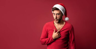 Kırmızı kazaklı ve Noel Baba şapkalı yakışıklı genç adam stüdyoda kırmızı arka planda duruyor ve ne kadar seksi olduğunu gösteriyor.