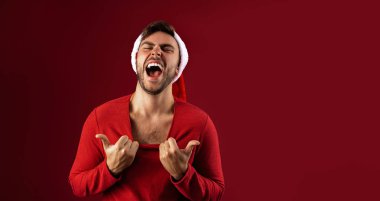 Kırmızı kazaklı ve Noel Baba şapkalı genç yakışıklı beyaz adam stüdyoda kırmızı arka planda duruyor. Ne kadar seksi olduğunu gösteriyor.