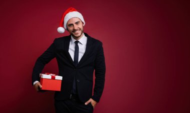 İş elbisesi içindeki yakışıklı beyaz adam ve Noel Baba şapkası stüdyoda kırmızı arka planda duruyor ve elinde kırmızı bir hediye kutusu tutuyor..