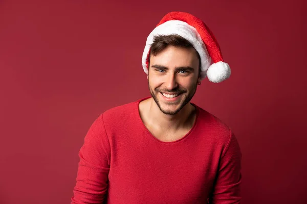 Jovem bonito caucasiano cara em camisola vermelha e chapéus de Papai Noel fica no fundo vermelho no estúdio e e dentes sorridentes — Fotografia de Stock