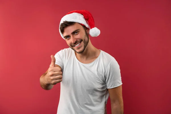 Jovem cara bonito caucasiano em uma camiseta branca e chapéus de Papai Noel fica no fundo vermelho no estúdio e mostrando os polegares para cima . — Fotografia de Stock