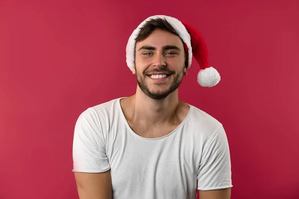 Jovem cara bonito caucasiano em uma camiseta branca e chapéus de Papai Noel fica no fundo vermelho no estúdio e e dentes sorridentes — Fotografia de Stock
