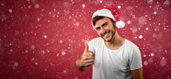 Jovem cara bonito caucasiano em uma camiseta branca e chapéus de Papai Noel fica no fundo vermelho no estúdio e mostrando os polegares para cima . — Fotografia de Stock