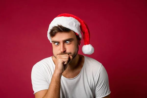 Jovem cara bonito caucasiano em uma camiseta branca e chapéus de Papai Noel fica no fundo vermelho no estúdio e doente após a comida de férias . — Fotografia de Stock