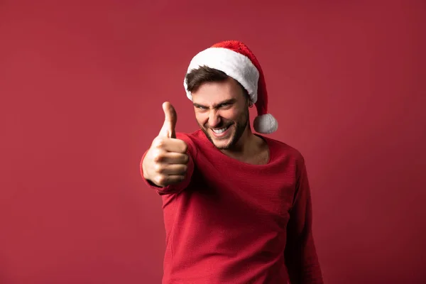 Junger gutaussehender kaukasischer Typ in rotem Pullover und Weihnachtsmannmütze steht auf rotem Hintergrund im Studio und zeigt Daumen hoch. — Stockfoto