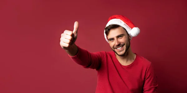 Jovem cara caucasiano bonito em uma camisola vermelha e chapéus de Papai Noel fica no fundo vermelho no estúdio e mostrando os polegares para cima . — Fotografia de Stock