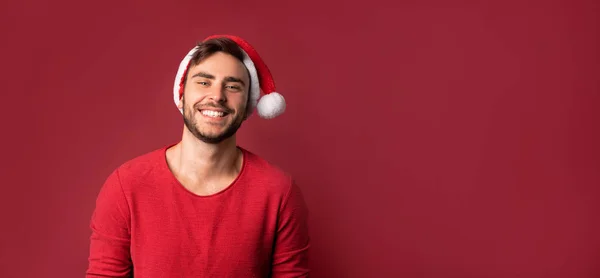 Junger gutaussehender kaukasischer Typ in rotem Pullover und Weihnachtsmannmütze steht auf rotem Hintergrund im Studio und lächelt — Stockfoto