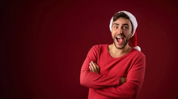 Junger gutaussehender kaukasischer Typ in rotem Pullover und Weihnachtsmannmütze steht auf rotem Hintergrund im Studio und lächelt — Stockfoto