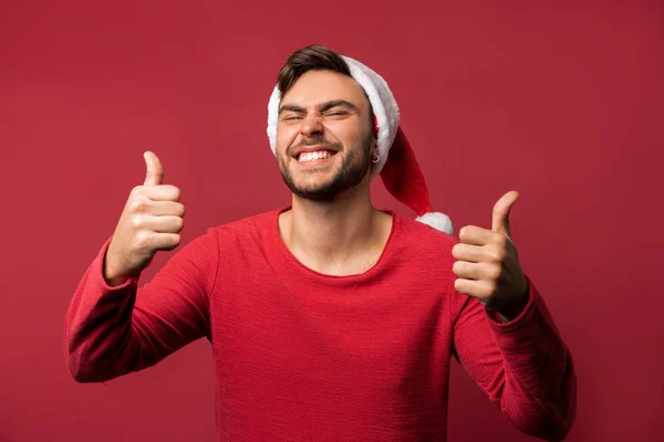 Jovem cara caucasiano bonito em uma camisola vermelha e chapéus de Papai Noel fica no fundo vermelho no estúdio e mostrando os polegares para cima . — Fotografia de Stock