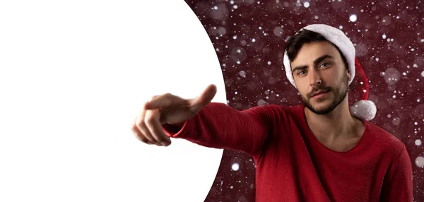 Junger gutaussehender kaukasischer Typ in rotem Pullover und Weihnachtsmannmütze steht auf rotem Hintergrund im Studio und zeigt den Finger in die Kamera. — Stockfoto