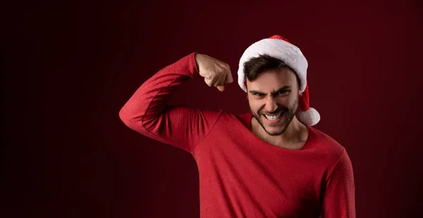 Jovem cara caucasiano bonito em uma camisola vermelha e chapéus de Papai Noel fica no fundo vermelho no estúdio e mostrando poder bíceps — Fotografia de Stock