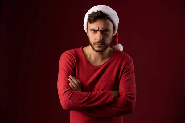 Junge ernsthafte gutaussehende kaukasische Kerl in rotem Pullover und Weihnachtsmützen steht auf rotem Hintergrund im Studio und ist wütend — Stockfoto