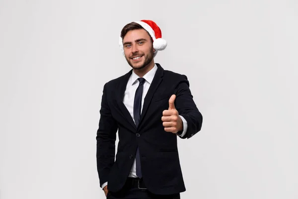 Jovem bonito caucasiano cara no negócio terno e Santa chapéus fica no branco fundo no estúdio smilie e mostrando polegares para cima . — Fotografia de Stock