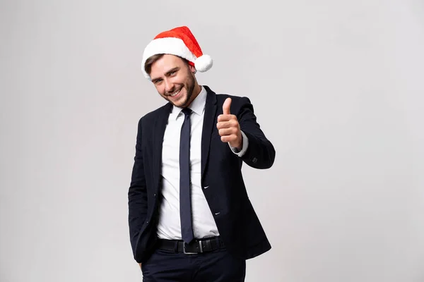 Jovem bonito caucasiano cara no negócio terno e Santa chapéus fica no branco fundo no estúdio smilie e mostrando polegares para cima . — Fotografia de Stock