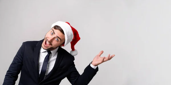 Jovem cara caucasiano bonito em terno de negócios e chapéus de Papai Noel fica no fundo branco em estúdio smilie e mostrando a mão para o lado . — Fotografia de Stock