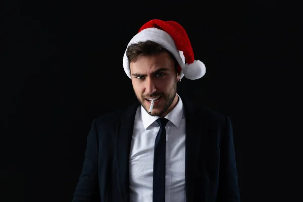 Young handsome caucasian guy in business suit and Santa hats stands on black background in studio with serious face and smoking cigarette — Stock Photo, Image