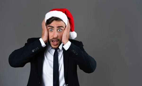 Jovem cara caucasiano bonito em terno de negócios e chapéus de Papai Noel fica em fundo cinza em estúdio mãos muito surpresas nas bochechas . — Fotografia de Stock