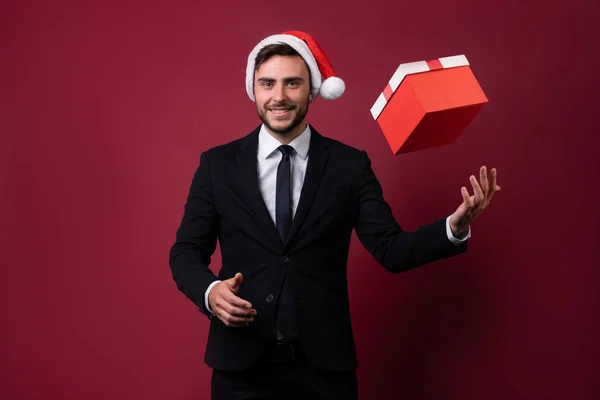 Jovem cara caucasiano bonito em terno de negócios e chapéus de Papai Noel fica no fundo vermelho no estúdio e smilie vomita caixa de presente vermelho na mão . — Fotografia de Stock