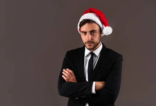 Jovem cara caucasiano bonito em terno de negócios e chapéus de Papai Noel fica em fundo cinza no estúdio com rosto sério braços dobrados no peito — Fotografia de Stock