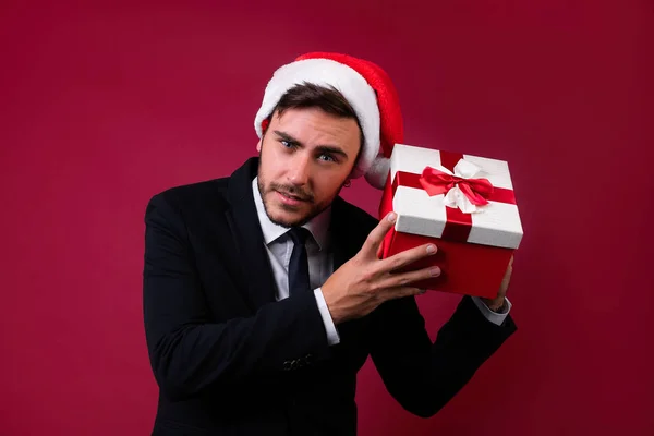 Jovem cara caucasiano bonito em terno de negócios e chapéus de Papai Noel fica no fundo vermelho no estúdio e smilie Holds perto da caixa de presente vermelho orelha — Fotografia de Stock