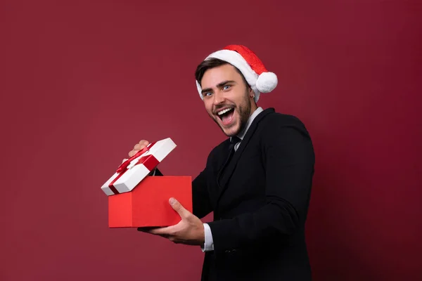 Junge hübsche kaukasische Kerl in Business-Anzug und Weihnachtsmützen steht auf rotem Hintergrund im Studio und Zähne smilie hält rote Geschenkbox in der Hand. — Stockfoto