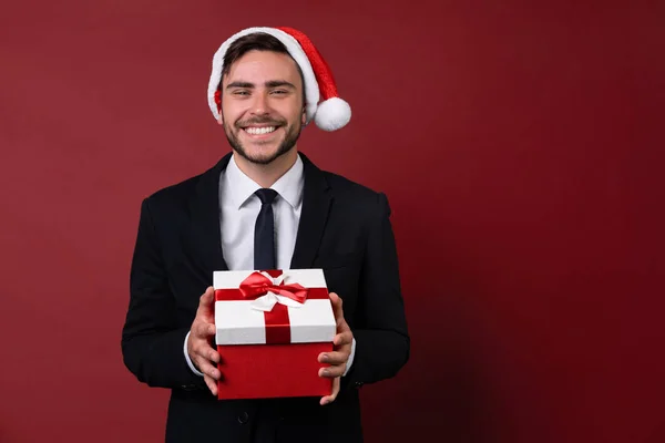 Jovem cara caucasiano bonito em terno de negócios e chapéus de Papai Noel fica no fundo vermelho no estúdio e dentes smilie Segurando caixa de presente vermelho na mão . — Fotografia de Stock
