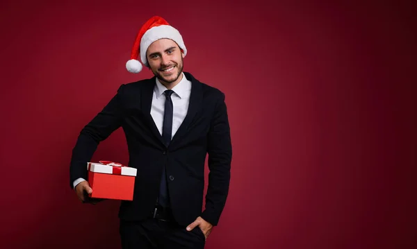 Jovem cara caucasiano bonito em terno de negócios e chapéus de Papai Noel fica no fundo vermelho no estúdio e smilie Segurando caixa de presente vermelho na mão . — Fotografia de Stock