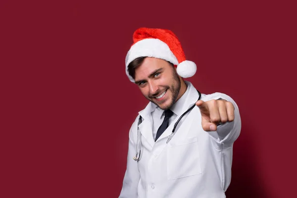 Jovem médico bonito em uniforme branco e chapéu de Papai Noel de pé em estúdio no fundo vermelho sorriso e dedo na câmera — Fotografia de Stock