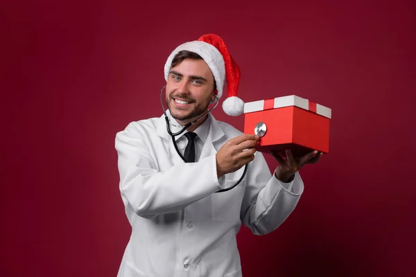 Jovem médico bonito em uniforme branco e chapéu de Papai Noel de pé em estúdio no fundo vermelho sorriso e dedo na câmera — Fotografia de Stock