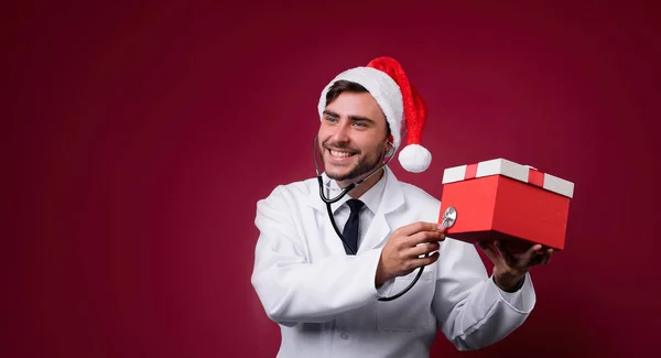 Beyaz üniformalı genç yakışıklı doktor ve Noel Baba şapkası stüdyoda kırmızı arka planda duruyor gülümseme ve kamera içinde parmak.
