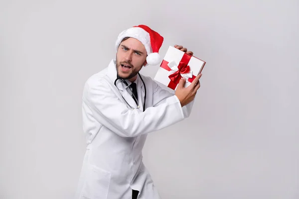 Joven médico guapo en uniforme blanco y sombrero de Santa Claus de pie en el estudio sobre fondo blanco sonrisa y el dedo en la cámara — Foto de Stock