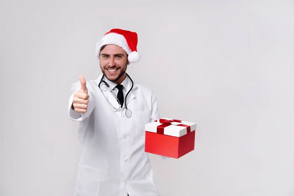 Beyaz üniformalı genç doktor ve Noel Baba şapkası stüdyoda beyaz arka planda gülümseme ve kamerada parmak.
