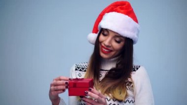 Genç, güzel, şehvetli Noel Baba kız beyaz kazak ve şirin Noel Baba şapkası giyip küçük kırmızı bir hediye kutusu açıyor. 