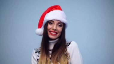 Beyaz kazaklı ve Noel Baba şapkalı genç ve güzel bir esmer. Stüdyoda mavi arka planda kameraya gülümsüyor..