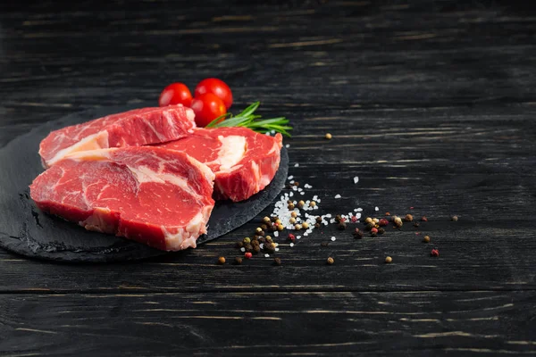 Trois morceaux de bœuf cru juteux sur une planche à découper en pierre sur un fond de table en bois noir . — Photo