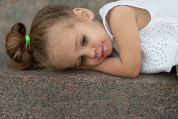 En vacker liten flicka med europeiskt utseende blont hår ligger ledsen på gatan asfalt. — Stockfoto