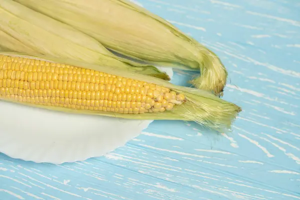 Corn cob with green leaves lies on white plate blue color background. — Stock Photo, Image