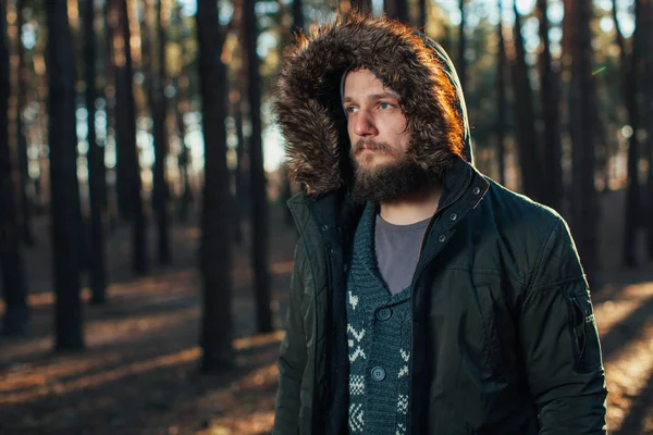 На фоне соснового леса стоит портрет молодого, стильно серьезного человека с бородой в зеленой зимней куртке с капюшоном и мехом на голове. . — стоковое фото