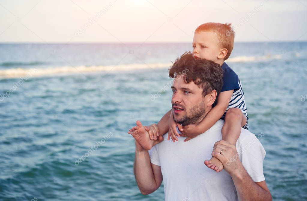 Young Caucasian dad with little son walk warm summer day along the sea coast. Summer family vacation concept. Friendship father and son. 