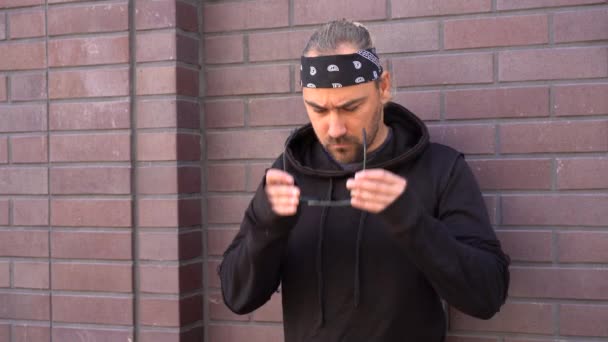 Handsome young unshaven man black hoodie and bandana stands backyard near brick walls summer day. — Stock Video