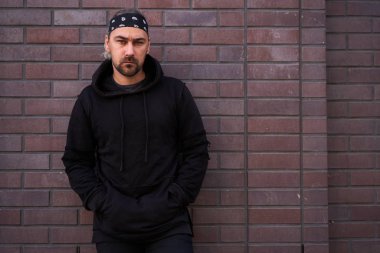 Yakışıklı, traşsız, siyah kapüşonlu ve bandanalı adam yaz günü tuğla duvarların yanında duruyor..