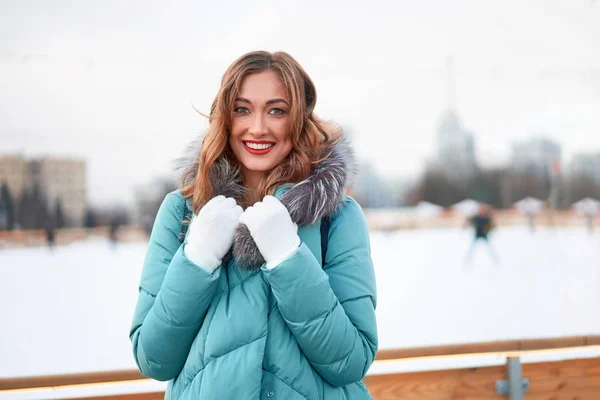 巻き毛の暖かい冬のジャケットを着た美しい中高年の女の子はアイスリンクの背景に立つ町の広場. — ストック写真