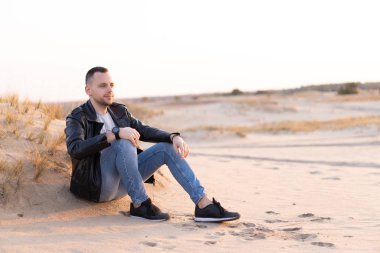 Siyah deri ceket ve kot pantolon giyen beyaz bir adam kumlu sahilde oturuyor. Yakışıklı Avrupalı adam Yalnız yürüyor